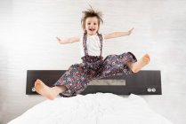 Content kid in overalls in moment of jumping on bed with outstretched arms and legs looking at camera — Stock Photo
