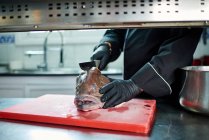 Unrecognizable cook in black uniform and gloves using metal cleaver while cutting head of large fresh fish on red chopped board at table against blurred interior of contemporary kitchen — Stock Photo