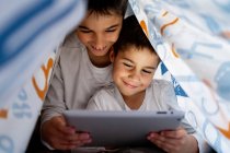 Hermanos positivos en pijama escondidos debajo de la manta y disfrutando de dibujos animados interesantes durante el día en casa - foto de stock
