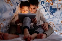 Позитивные братья и сестры в пижаме прячутся под одеялом и наслаждаются интересным мультфильмом в дневное время дома — стоковое фото