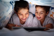 Frères et sœurs positifs en pyjama se cachant sous la couverture et appréciant un dessin animé intéressant pendant la journée à la maison — Photo de stock