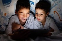 Irmãos positivos em pijamas escondidos sob cobertor e desfrutando de desenhos animados interessantes durante o dia em casa — Fotografia de Stock