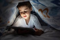 Adorable niño en pijama escondido debajo de la manta y el uso de la tableta mientras se entretiene durante la noche en casa - foto de stock