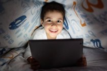 Adorabile bambino in pigiama nascosto sotto la coperta e utilizzando tablet durante il divertimento durante la notte a casa — Foto stock