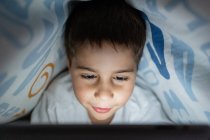 Liebenswertes Kind im Schlafanzug versteckt sich unter einer Decke und benutzt Tabletten, während es nachts zu Hause unterhält — Stockfoto