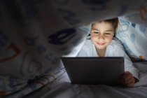 Criança adorável em pijama escondido sob cobertor e usando tablet enquanto se diverte durante a noite em casa — Fotografia de Stock