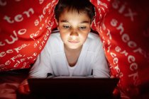Adorabile bambino in pigiama nascosto sotto la coperta e utilizzando tablet durante il divertimento durante la notte a casa — Foto stock