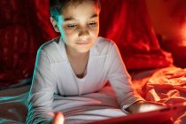 Adorable kid in pajamas hiding under blanket and using tablet while entertaining during night at home — Stock Photo