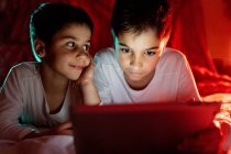 Positive Geschwister im Pyjama, die sich unter einer Decke verstecken und tagsüber zu Hause interessante Zeichentrickfilme genießen — Stockfoto