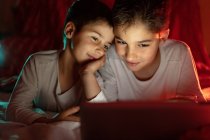 Positive siblings in pajamas hiding under blanket and enjoying interesting cartoon during daytime at home — Stock Photo