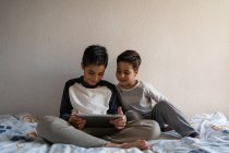 Sorridenti fratelli in pigiama party seduti su un letto accogliente e guardare film insieme mentre si divertono a casa — Foto stock