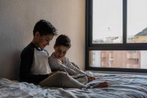 Irmãos sorridentes em roupas de dormir sentados na cama aconchegante e assistindo filme juntos enquanto entretendo em casa — Fotografia de Stock