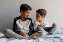 Irmãos sorridentes em roupas de dormir sentados na cama aconchegante e assistindo filme juntos enquanto entretendo em casa — Fotografia de Stock