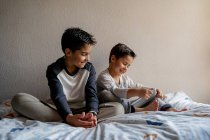 Sonrientes hermanos en ropa de dormir sentados en una cama acogedora y viendo películas juntos mientras se entretienen en casa - foto de stock