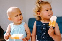Милые маленькие девочки едят вкусное мороженое, наслаждаясь летом и сидя вместе на заднем дворе — стоковое фото