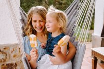 Веселая мать и дочь обнимаются в гамаке на террасе с вкусными ледяными леденцами и наслаждаются летом вместе — стоковое фото