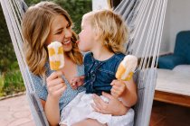 Cheerful mother and daughter hugging in hammock on terrace with tasty ice lollies and enjoying summer together — Stock Photo