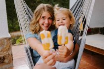 Весела мати і дочка обіймаються в гамаку на терасі зі смачними льодовиками і насолоджуються літом разом, дивлячись на камеру — стокове фото