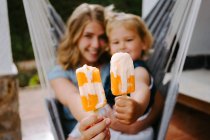 Cheerful mother and daughter hugging in hammock on terrace with tasty ice lollies and enjoying summer together looking at camera — Stock Photo