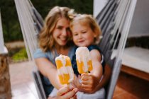 Веселая мать и дочь обнимаются в гамаке на террасе с вкусными ледяными леденцами и наслаждаются летом вместе, глядя в камеру — стоковое фото