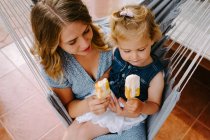 Fröhliche Mutter und Tochter umarmen sich in der Hängematte auf der Terrasse mit leckeren Eislutschern und genießen den Sommer zusammen — Stockfoto