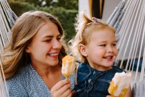 Веселая мать и дочь обнимаются в гамаке на террасе с вкусными ледяными леденцами и наслаждаются летом вместе — стоковое фото