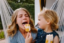 Allegro mamma e figlia che si abbracciano in amaca sulla terrazza con gustosi lecca-lecca e godersi l'estate insieme — Foto stock