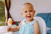 Симпатичный малыш, сидящий на высоком стуле на террасе и поедающий вкусное мороженое, глядя в сторону — стоковое фото