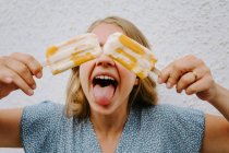 Feminino fazendo careta engraçada com língua para fora e cobrindo os olhos com saborosos piruetas de gelo em paus — Fotografia de Stock