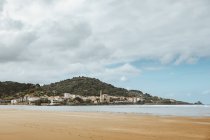 Спокойный океан с песчаным побережьем рядом с зеленой горой со зданиями под облачным небом — стоковое фото
