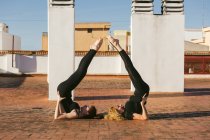 Side view of young female with mature mother doing Supported Shoulder Stand together while practicing partner yoga on rooftop of city building in sunny day — Stock Photo