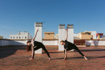 Ganzkörper-Seitenansicht schlanker Frauen in schwarzer Aktivkleidung, die Revolved Triangle Pose beim gemeinsamen Yoga auf der Dachterrasse der Stadt ausführen — Stockfoto
