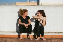 Fröhliche junge Frau mit Mutter mittleren Alters in Sportkleidung plaudert und lacht, während sie mit einer Flasche Wasser auf dem Dach sitzt und sich nach dem gemeinsamen Yoga ausruht — Stockfoto