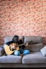 Mulher tocando guitarra sentada em seu sofá em casa e aprendendo com aulas on-line e algumas máscaras estão penduradas devido à contenção. Atrás dele está uma parede de tijolo — Fotografia de Stock