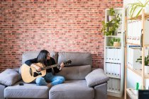 Жінка, яка грає на гітарі, сидить на дивані вдома і вчиться на онлайн уроках, а деякі маски звисають через стримування. За нею цегляна стіна — стокове фото