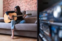 Woman playing guitar sitting on her couch at home and learning with online lessons and some masks are hanging due to containment. Behind it is a brick wall — Stock Photo