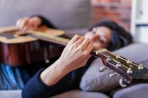 Mulher tocando guitarra sentada em seu sofá em casa e aprendendo com aulas on-line e algumas máscaras estão penduradas devido à contenção. Atrás dele está uma parede de tijolo — Fotografia de Stock
