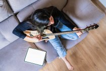 Mujer tocando la guitarra sentada en su sofá en casa y aprendiendo con lecciones en línea con una tableta digital con una tableta digital con una pantalla en blanco desde arriba - foto de stock