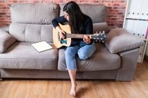 Mulher tocando guitarra sentada em seu sofá em casa e aprendendo com aulas on-line com um tablet digital com um tablet digital com uma tela em branco de cima — Fotografia de Stock