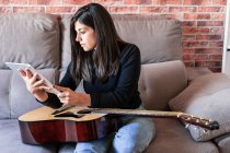 Жінка грає на гітарі, сидить на дивані вдома і вчиться з онлайн-уроками з цифровим планшетом з цифровим планшетом з порожнім екраном зверху — стокове фото