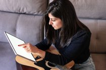 Frau, die zu Hause auf ihrer Couch Gitarre spielt und mit einem digitalen Tablet mit einem leeren Bildschirm von oben mit Online-Unterricht lernt — Stockfoto