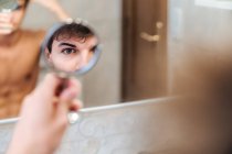 Sério masculino em pé no banheiro brilhante e olhando em espelho redondo de manhã — Fotografia de Stock
