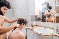 Male with hair trimmer cutting hair of guy in contemporary bathroom at home — Stock Photo