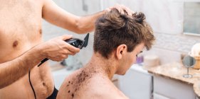 Male with hair trimmer cutting hair of guy in contemporary bathroom at home — Stock Photo