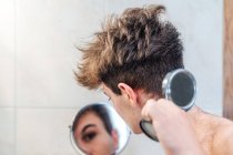 Спокойный парень с обнаженным туловищем, смотрящий на себя в дополнительное маленькое зеркало после самообрезки волос в светлой ванной комнате дома — стоковое фото