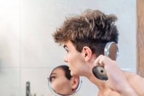 Ruhiger Typ mit nacktem Oberkörper, der sich selbst in einem zusätzlichen kleinen Spiegel ansieht, nachdem er sich die Haare im hellen Badezimmer zu Hause abgeschnitten hat — Stockfoto