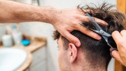 Back view of unrecognizable male making haircut to guy using scissors against blurred interior of light bathroom at home — Stock Photo