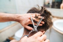 Back view of unrecognizable male making haircut to guy using scissors against blurred interior of light bathroom at home — Stock Photo