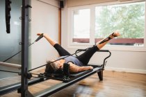 Femme adulte en vêtements de sport couchée sur pilates machine et étirant les jambes avec des bandes de résistance pendant l'entraînement dans la salle de gym moderne — Photo de stock