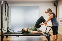 Concentrated male in sportswear doing exercises on pilates reformer during training under supervision of female instructor in protective respirator during coronavirus pandemic — Stock Photo